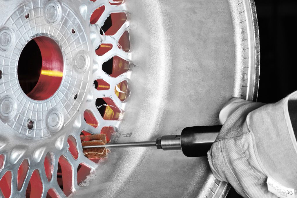 A miniature mounted flap wheel grinds the edges of a broken aluminum rim. A person wearing protective gloves guides the tool.